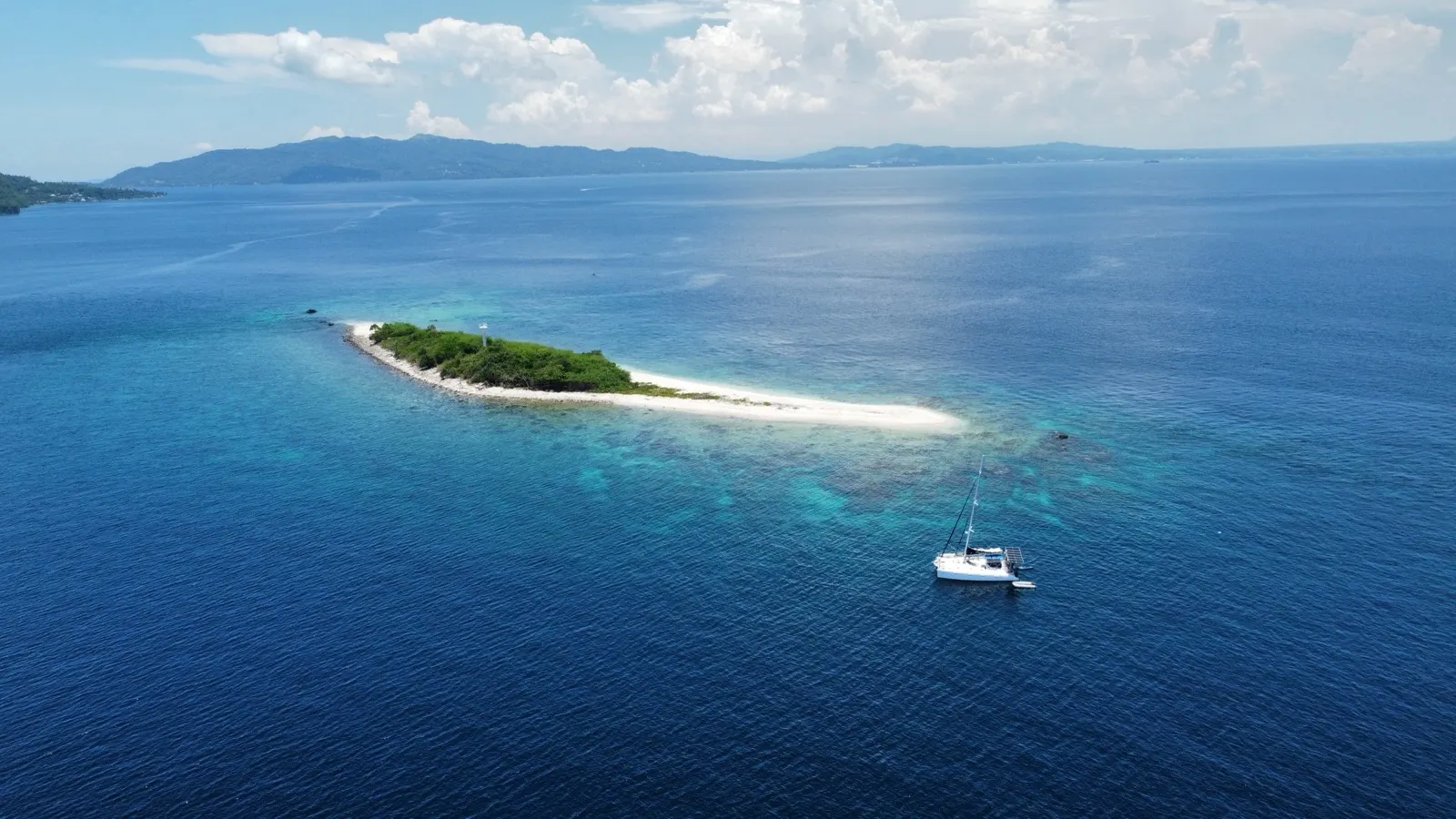Wide aerial of sandbar island with catamaran anchored, coastline behind