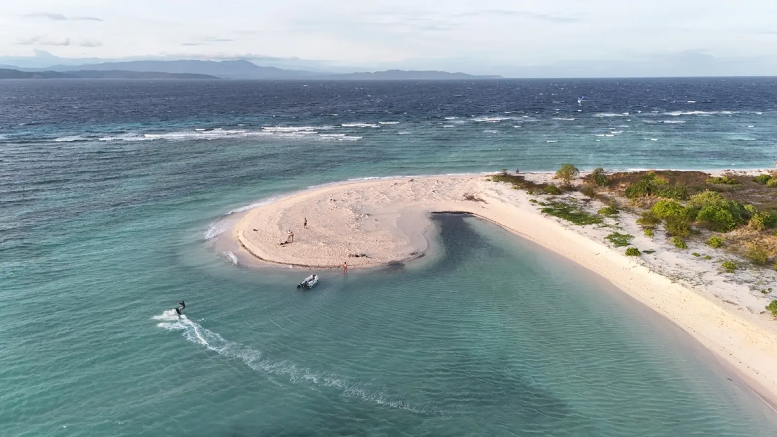 Kitesurfer jumping near a sandbar, dinghy and people nearby