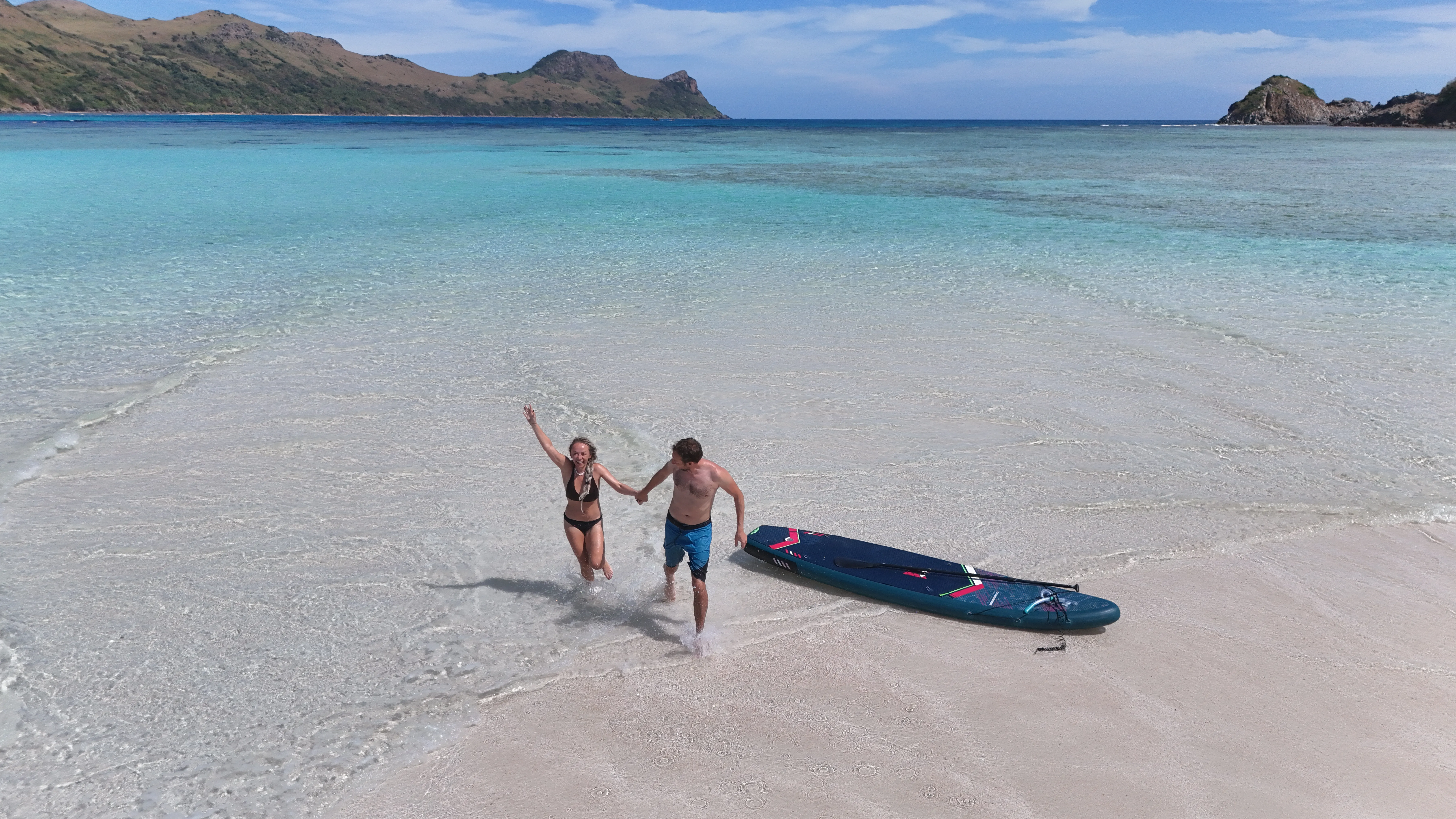 A couple running up a beach with a paddleboard