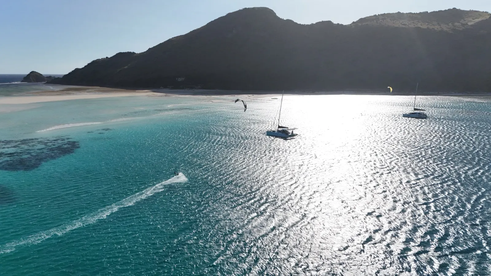 Kitesurfers and catamarans backlit near beach in sun glare