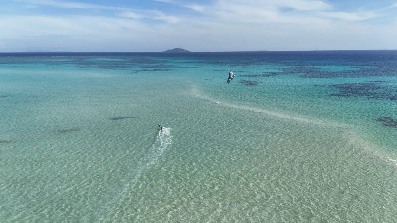 Distant aerial of a kitesurfer crossing a pale lagoon