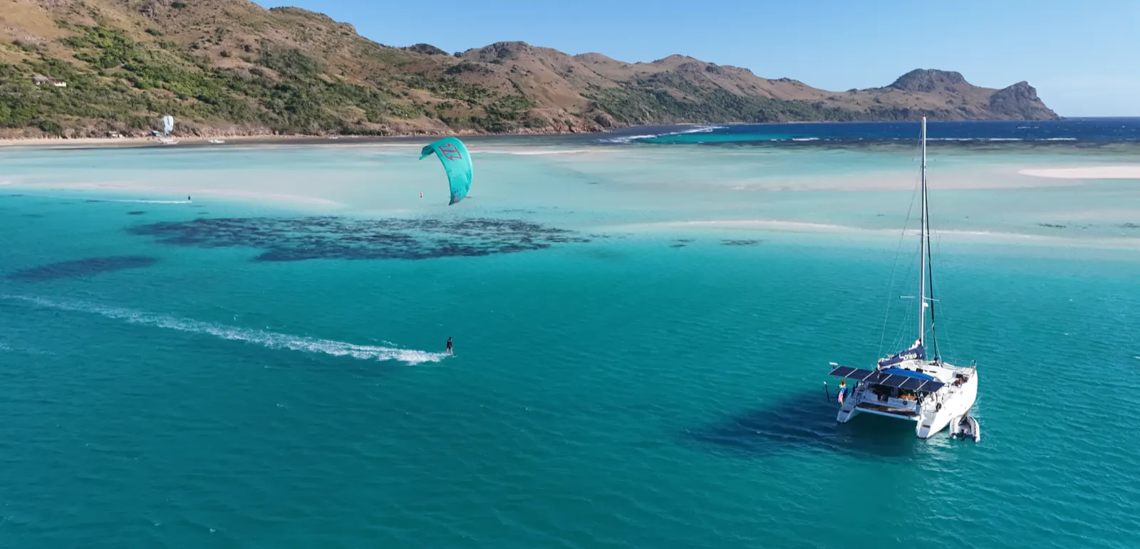 Catamaran anchored off mountainous coastline, kitesurfer nearby