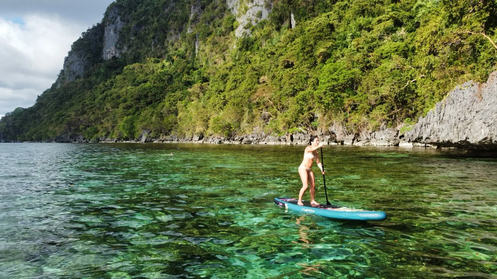 Woman paddleboarding through clear water beneath limestone cliffs
