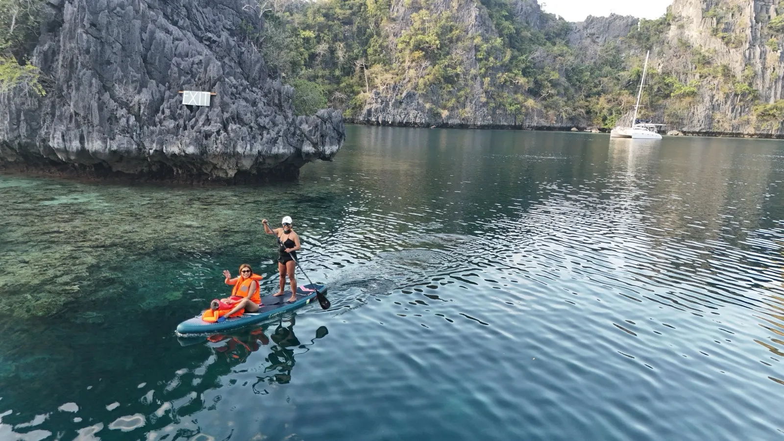 Two women on paddleboard in limestone cove, catamaran behind