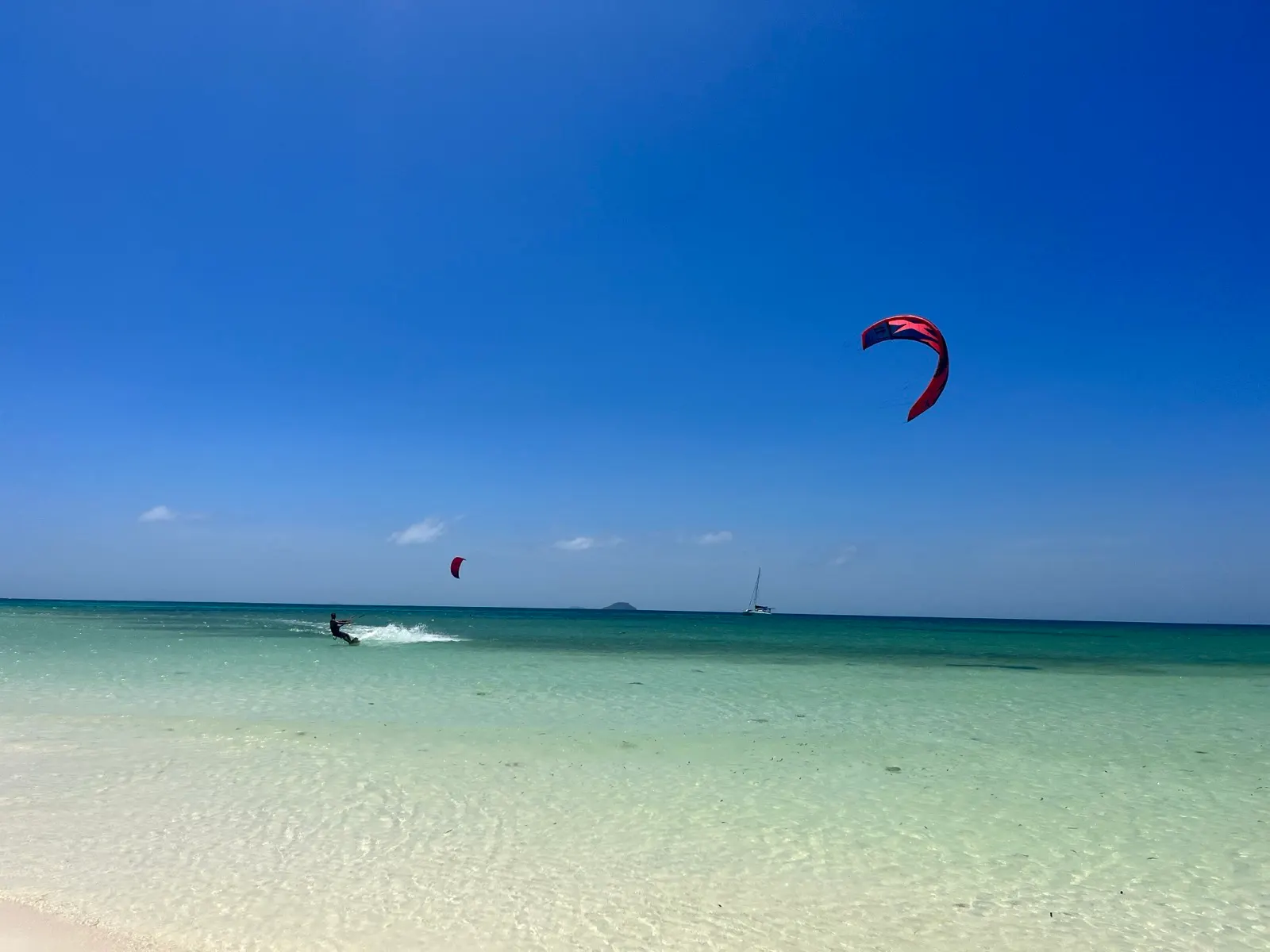 Kitesurfer riding turquoise shallows near sandbar, sailboat distant
