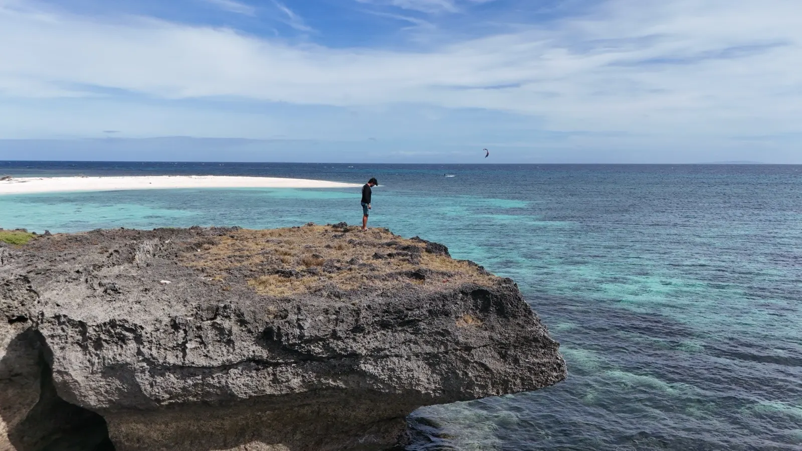 Man on rock outcrop overlooking ocean, kitesurfer distant