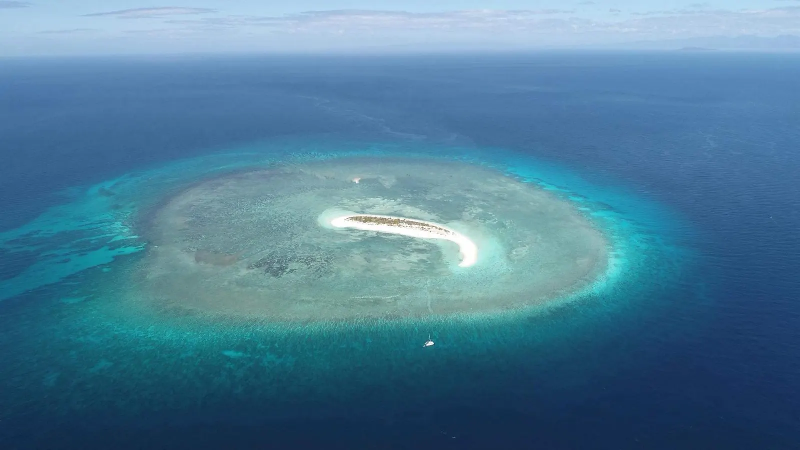 Aerial of tiny sand island ringed by reef, boat anchored