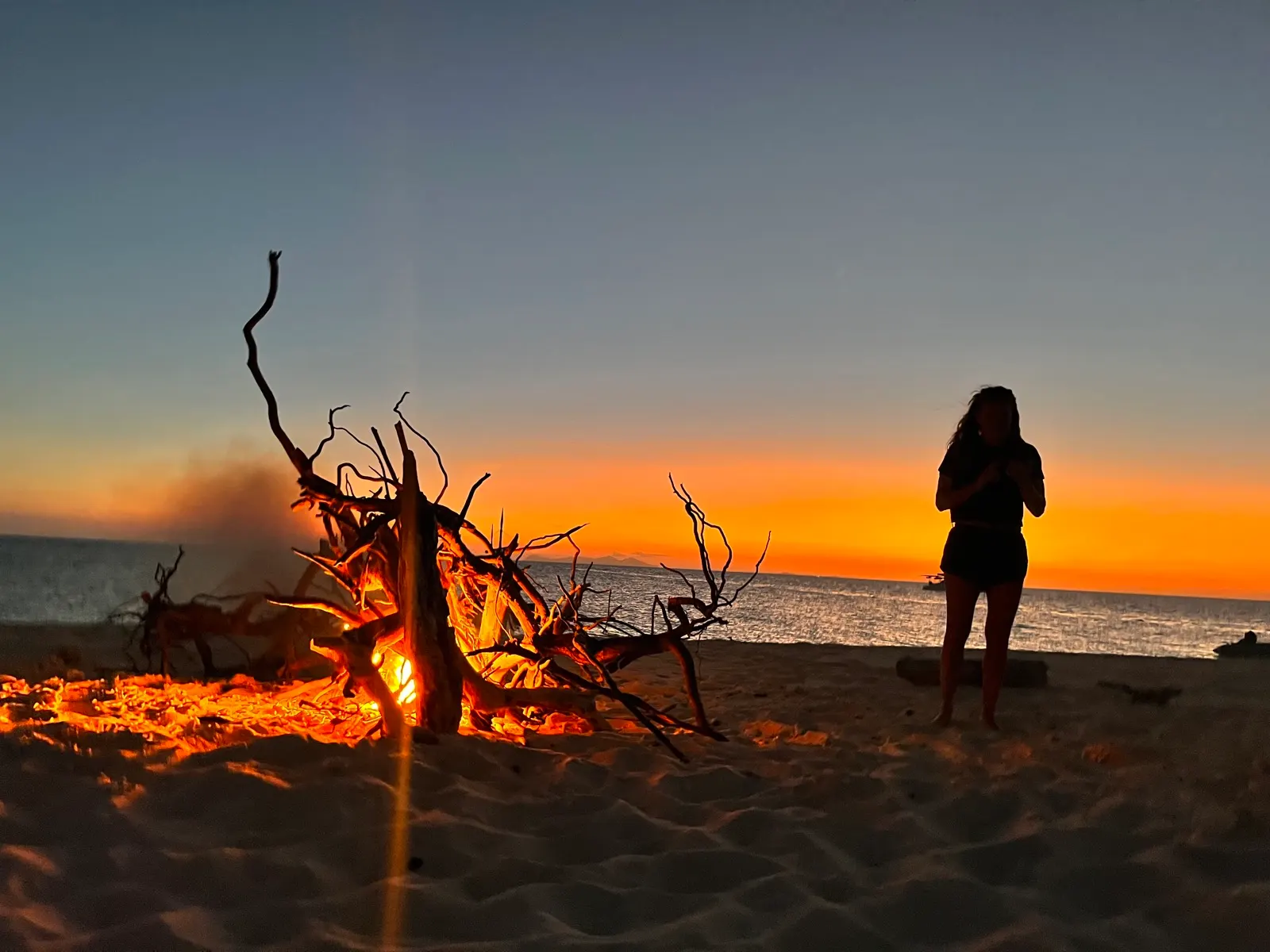 Silhouetted woman beside beach bonfire at sunset